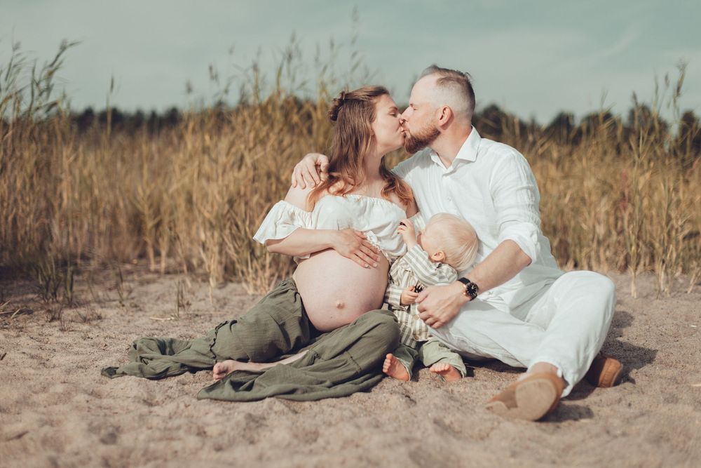 Couple kissing on sandy beach with pregnant belly and toddler sitting between them near tall grass. Perhekuvaus, odotuskuvaus.