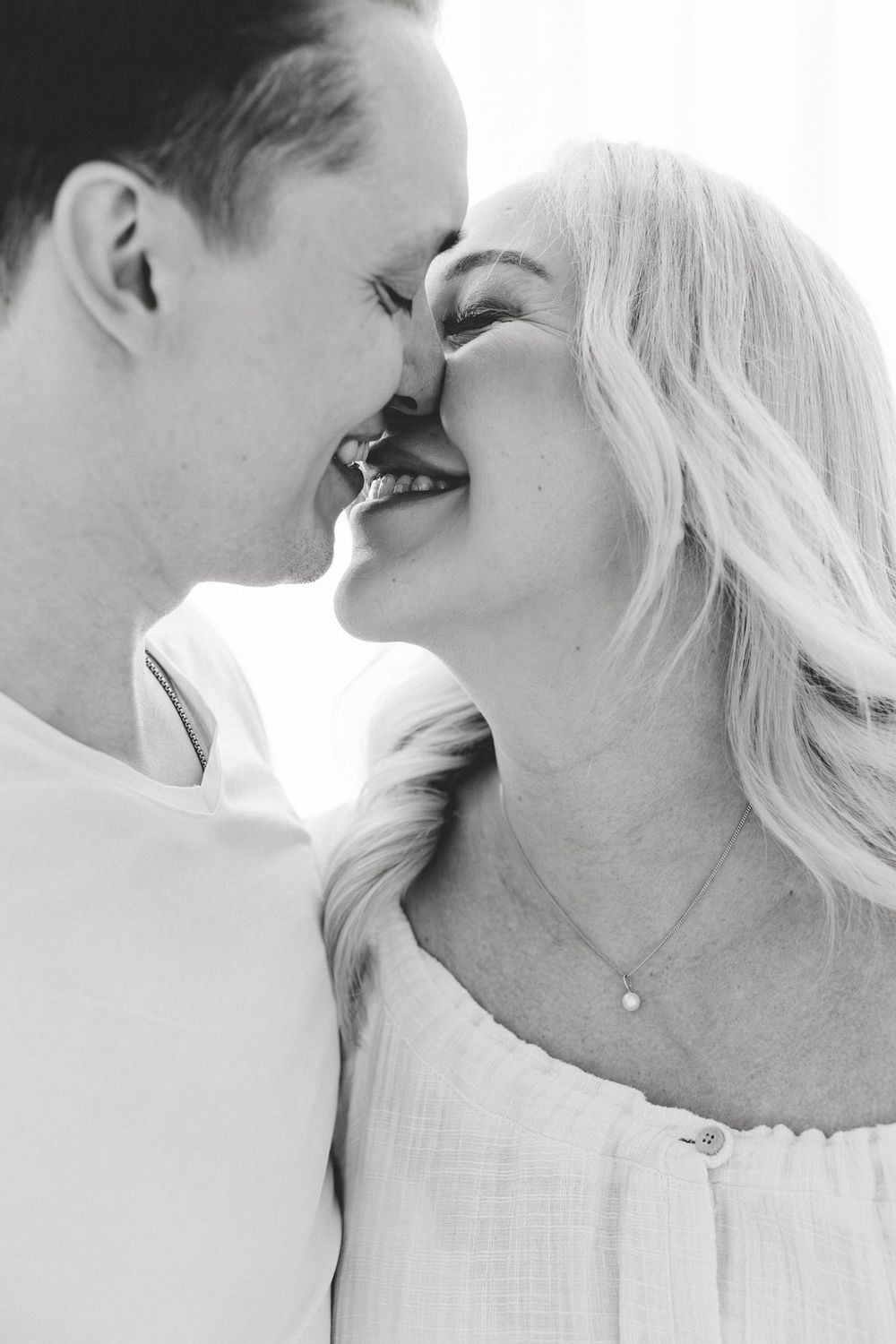 Black and white photo of a smiling couple leaning in for a kiss, woman wearing a delicate necklace.