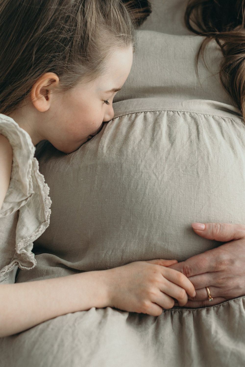 A young girl tenderly rests her head on her pregnant mother's belly, both wearing neutral linen clothing.