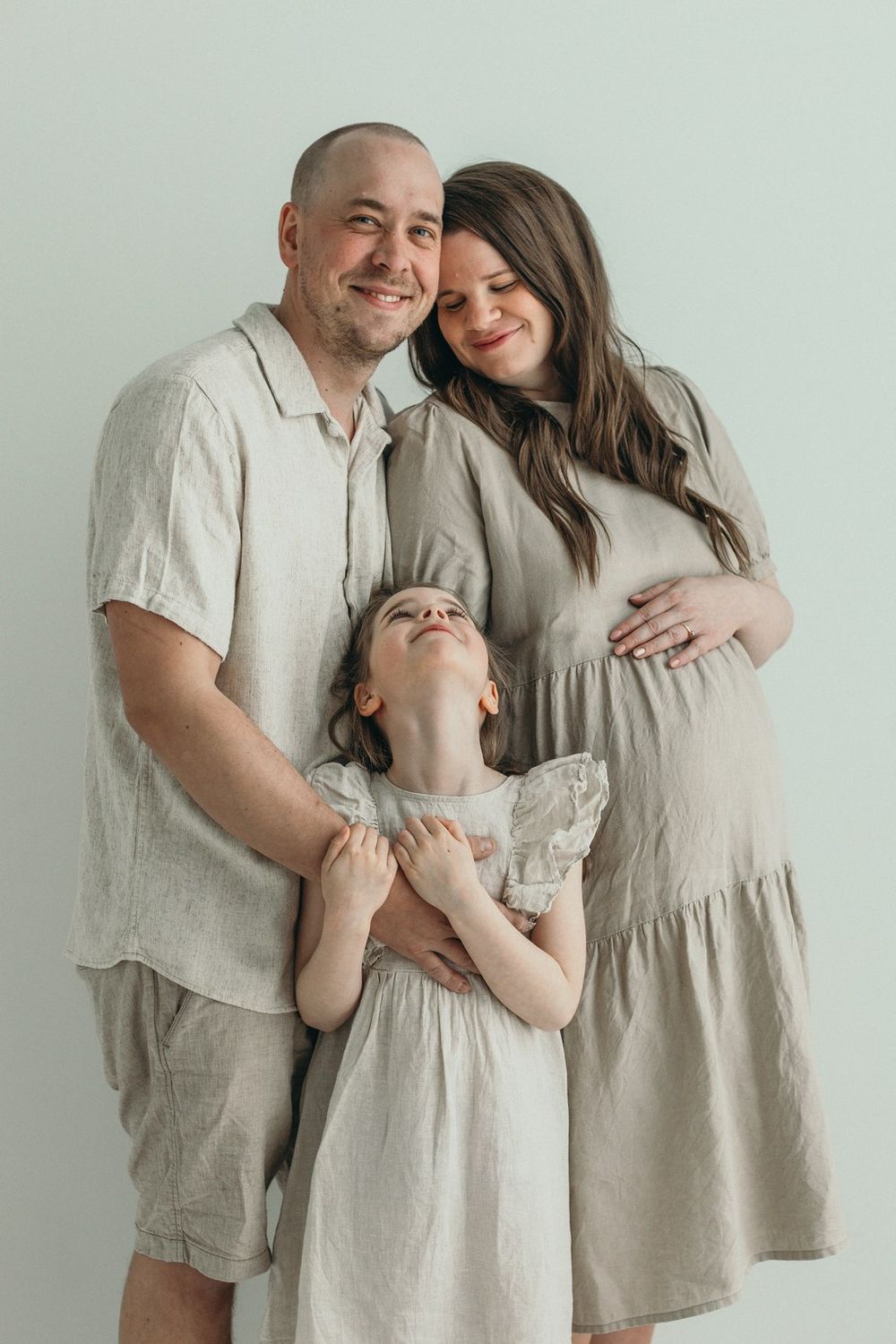 A smiling family of three in matching beige linen outfits, with a pregnant mother, father, and young daughter posing together.