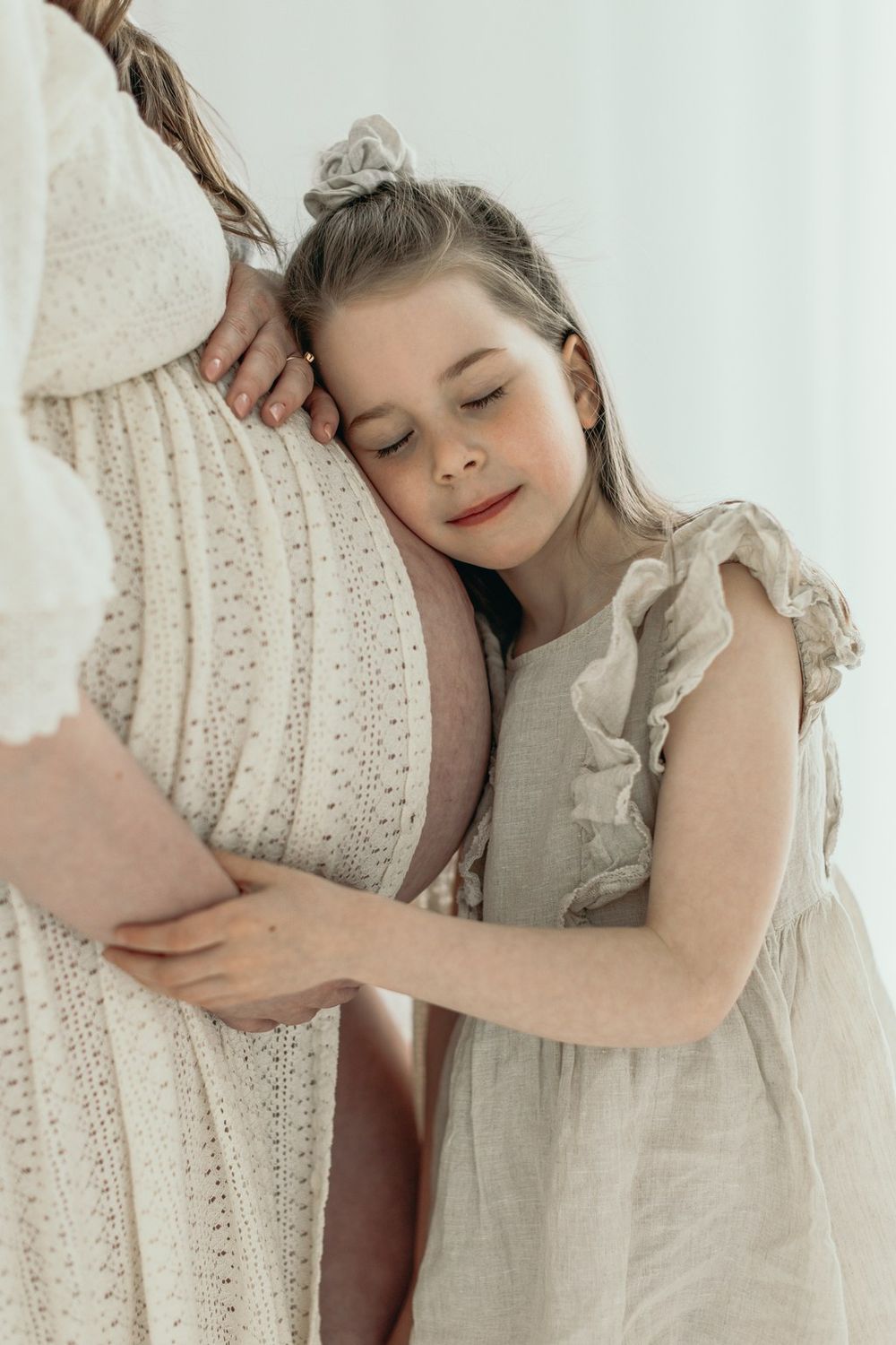 A young girl in a linen dress tenderly hugs her pregnant mother's belly with eyes closed.