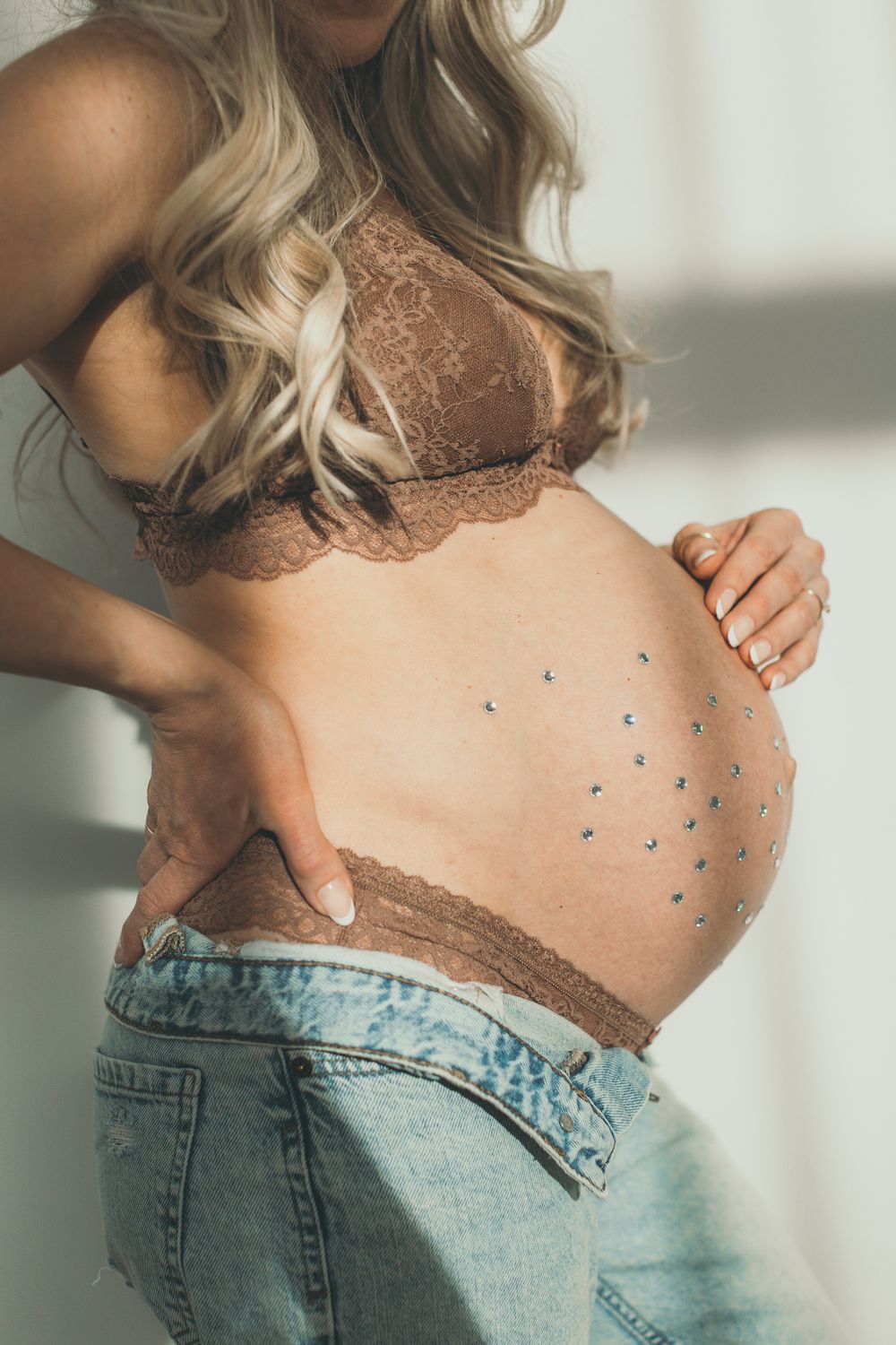 Pregnant woman in brown lace bralette and jeans with gemstone belly art highlighting her baby bump.