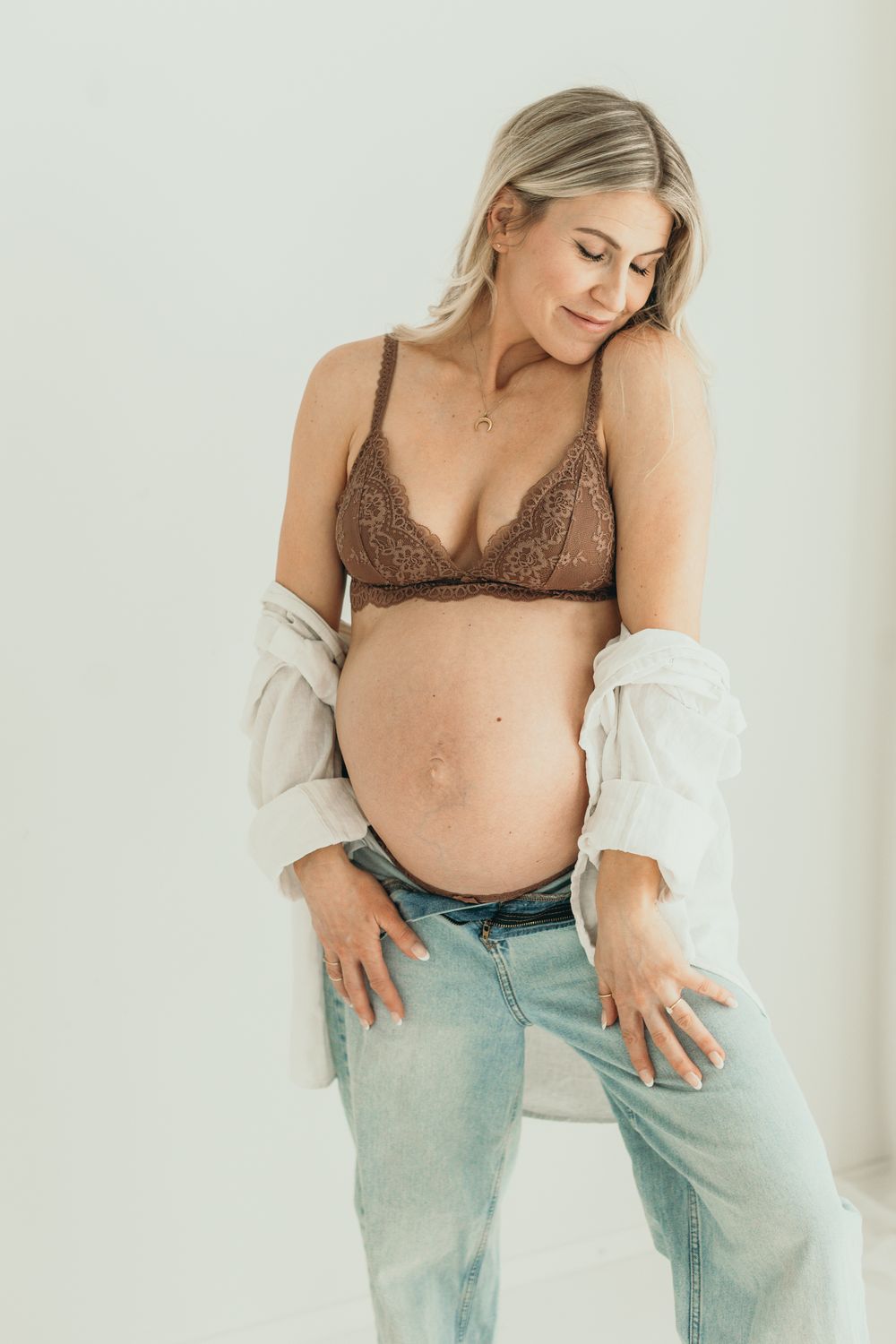 Pregnant woman in brown lace bralette and open white shirt cradles her baby bump while seated.