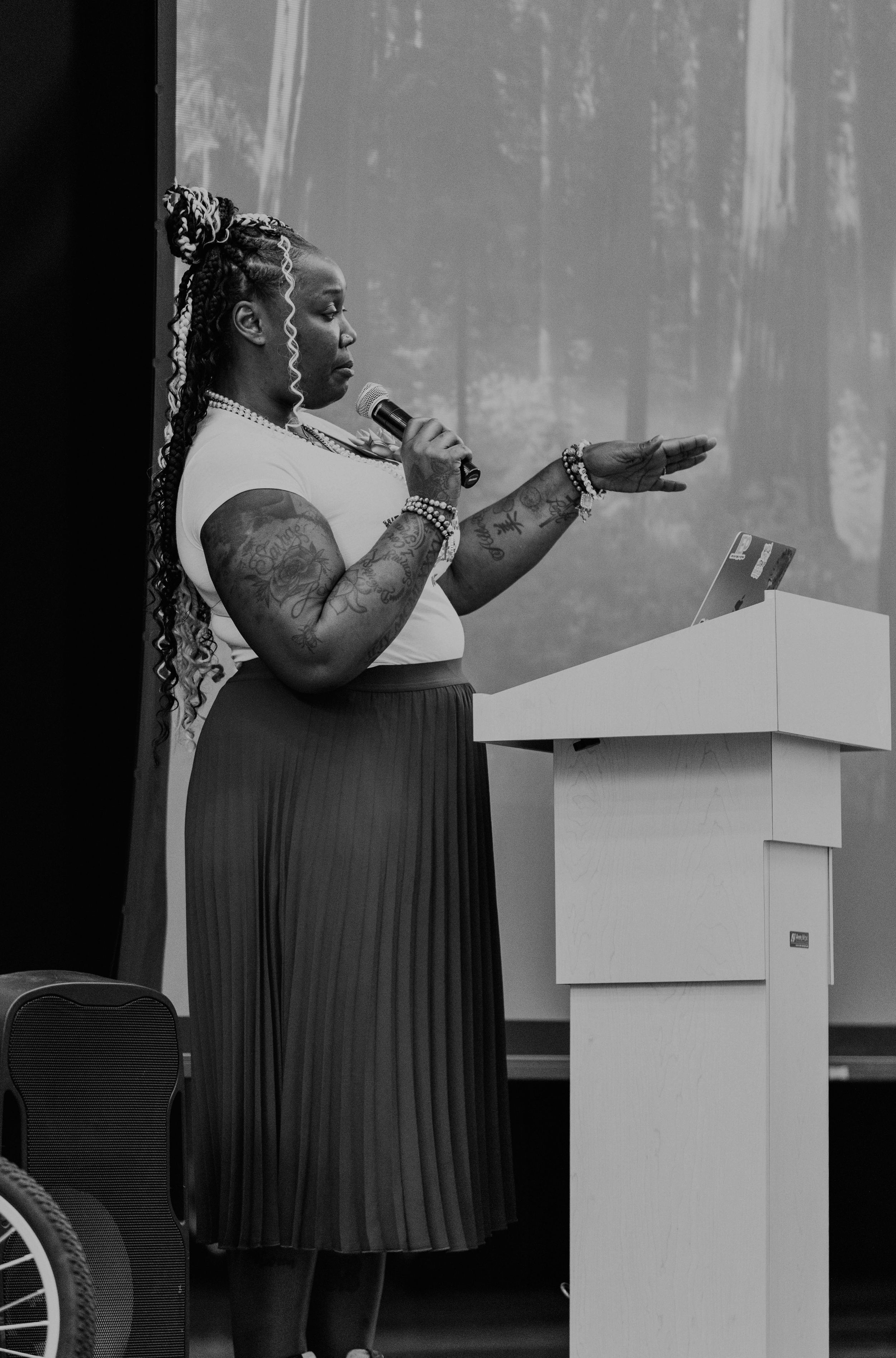 A speaker in a pleated dress gestures while presenting at a podium.