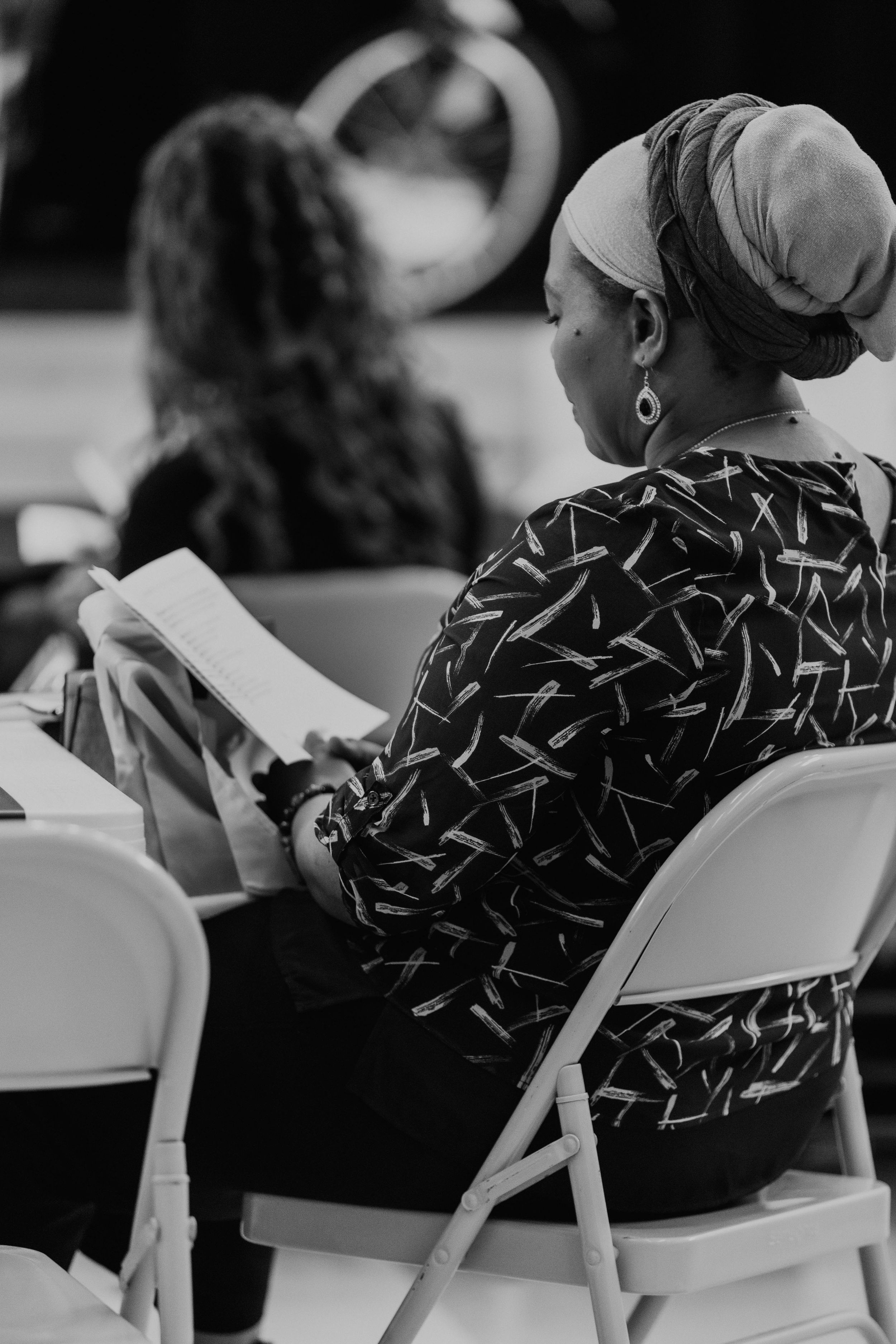 A person in a patterned top and headwrap reads from a book while seated in a folding chair.