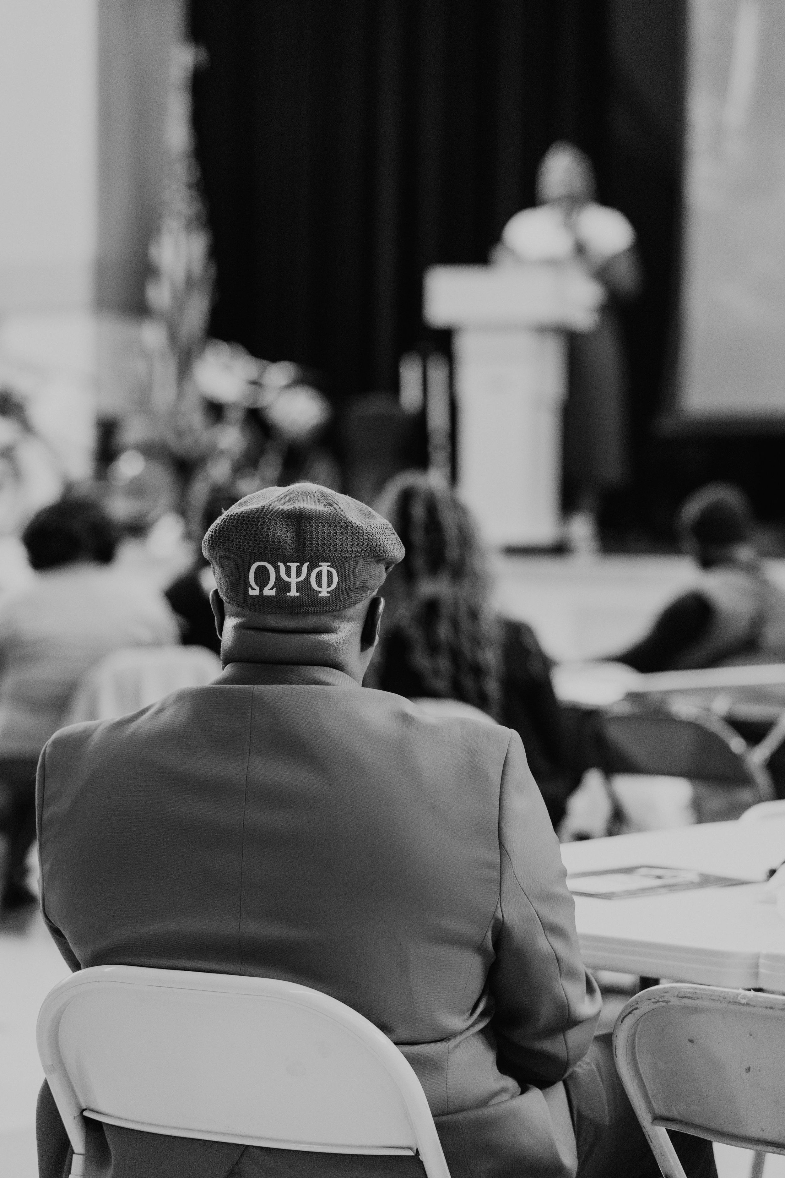 An attendee wearing a Greek letter cap watches a presentation in an auditorium.