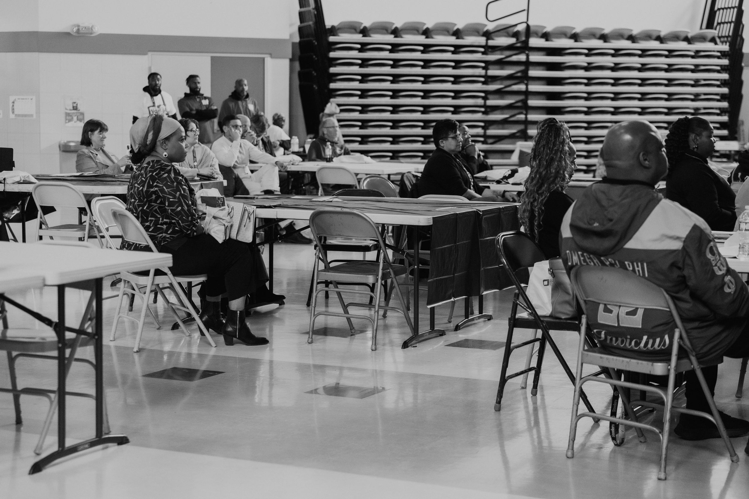 People seated at folding tables in a community hall with stacked chairs visible in background.