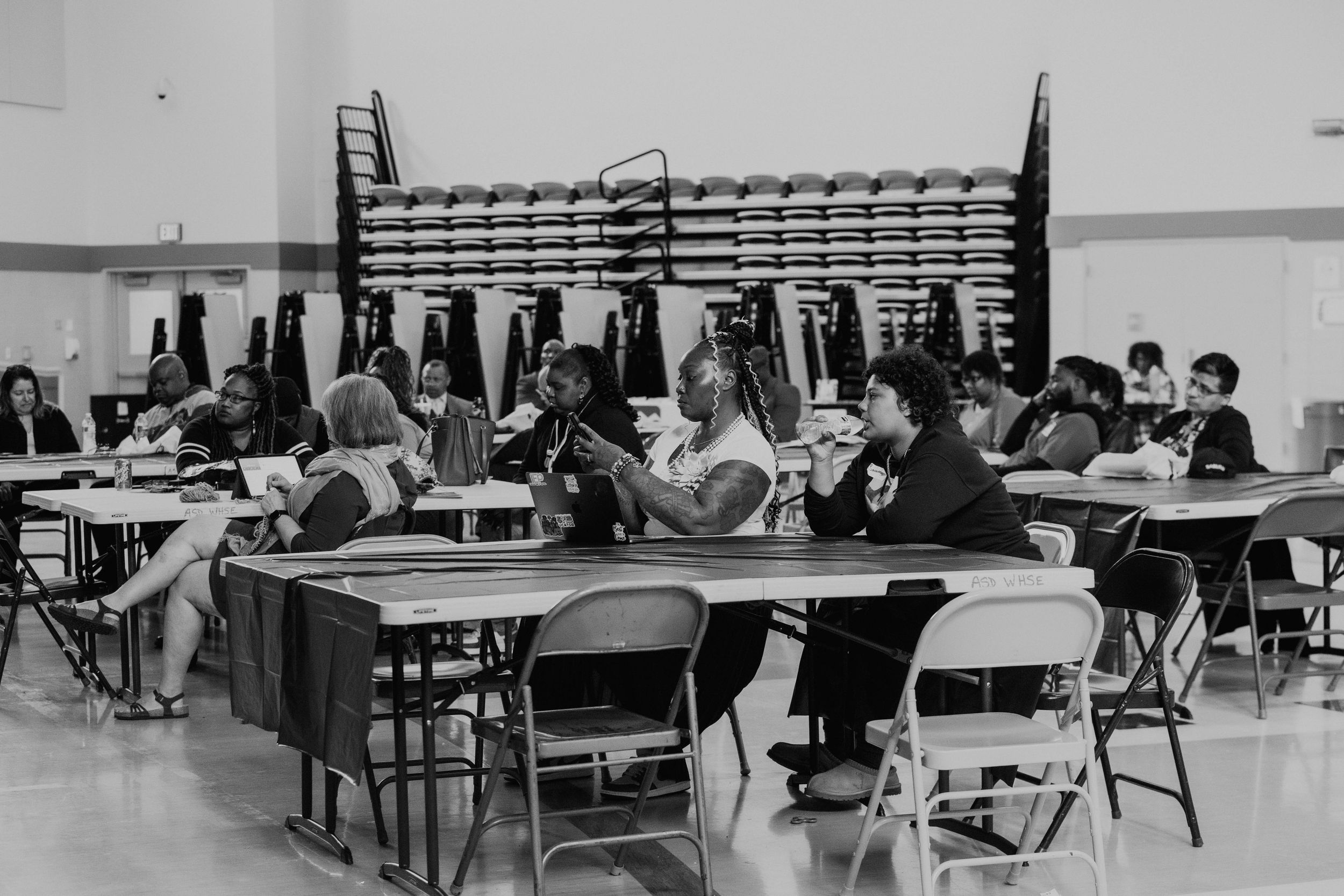 Students sitting at tables in a cafeteria or meeting room with folding chairs.