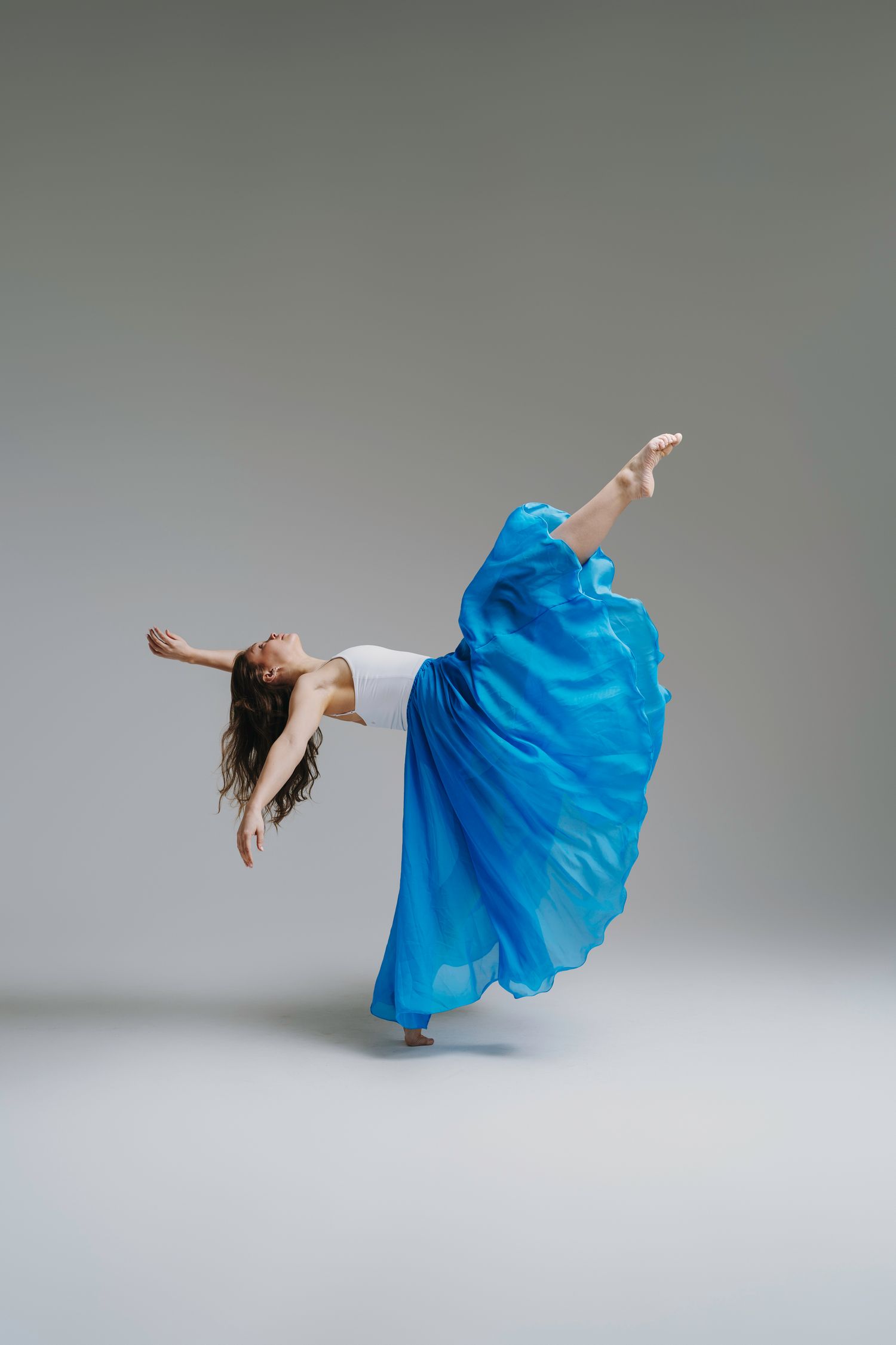 A dancer in a flowing bright blue dress performs an expressive movement sequence against a neutral gray backdrop.