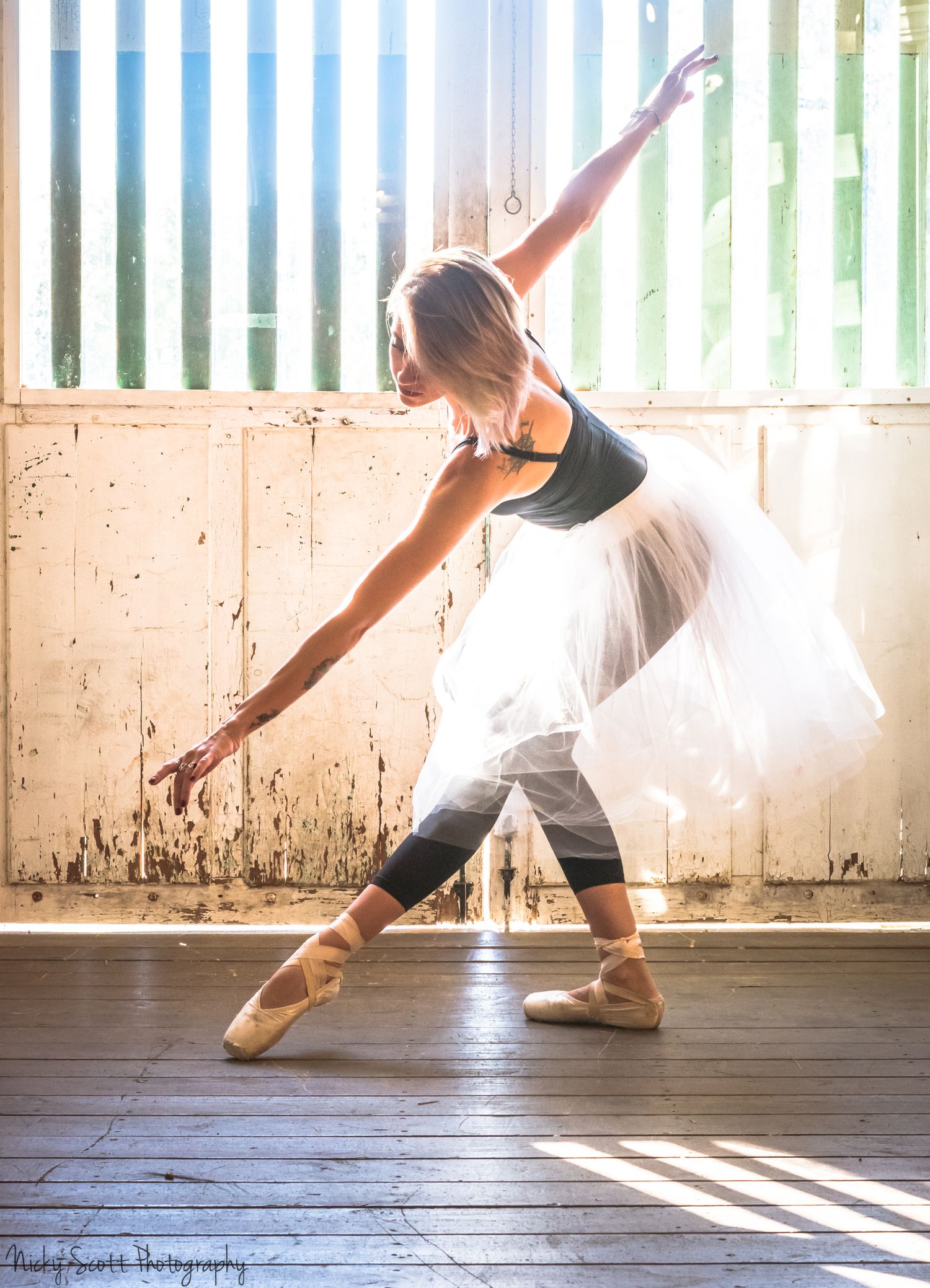Ballet dancer in tutu and leggings strikes graceful pose in sunlit studio.