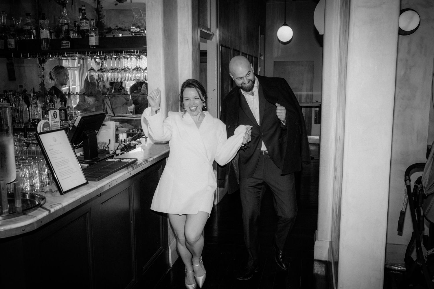 A black and white photograph shows guests dancing and laughing at a bar with wine glasses visible in the background.