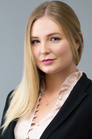 Professional headshot of someone with long blonde hair wearing a black blazer and pink blouse against a gray background.