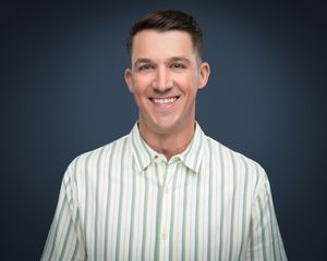 A confident person wearing a white and green striped button-up shirt smiles against a dark studio background.