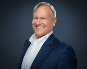 Professional headshot of someone in a navy blazer and white dress shirt smiling warmly against a dark background.