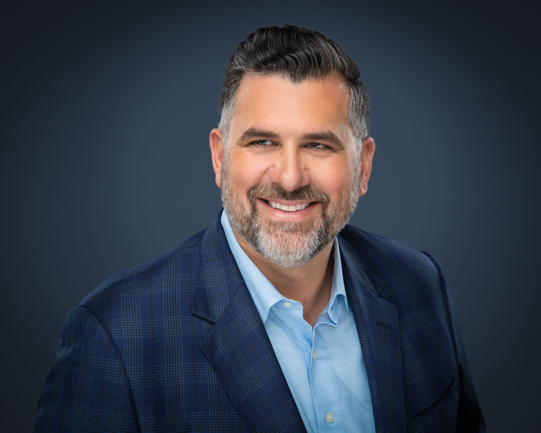 Professional headshot of person in navy plaid suit and light blue dress shirt against dark background.