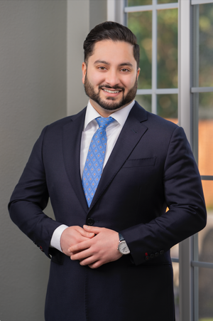 Professional headshot of someone in a dark suit and blue patterned tie standing in front of French doors.
