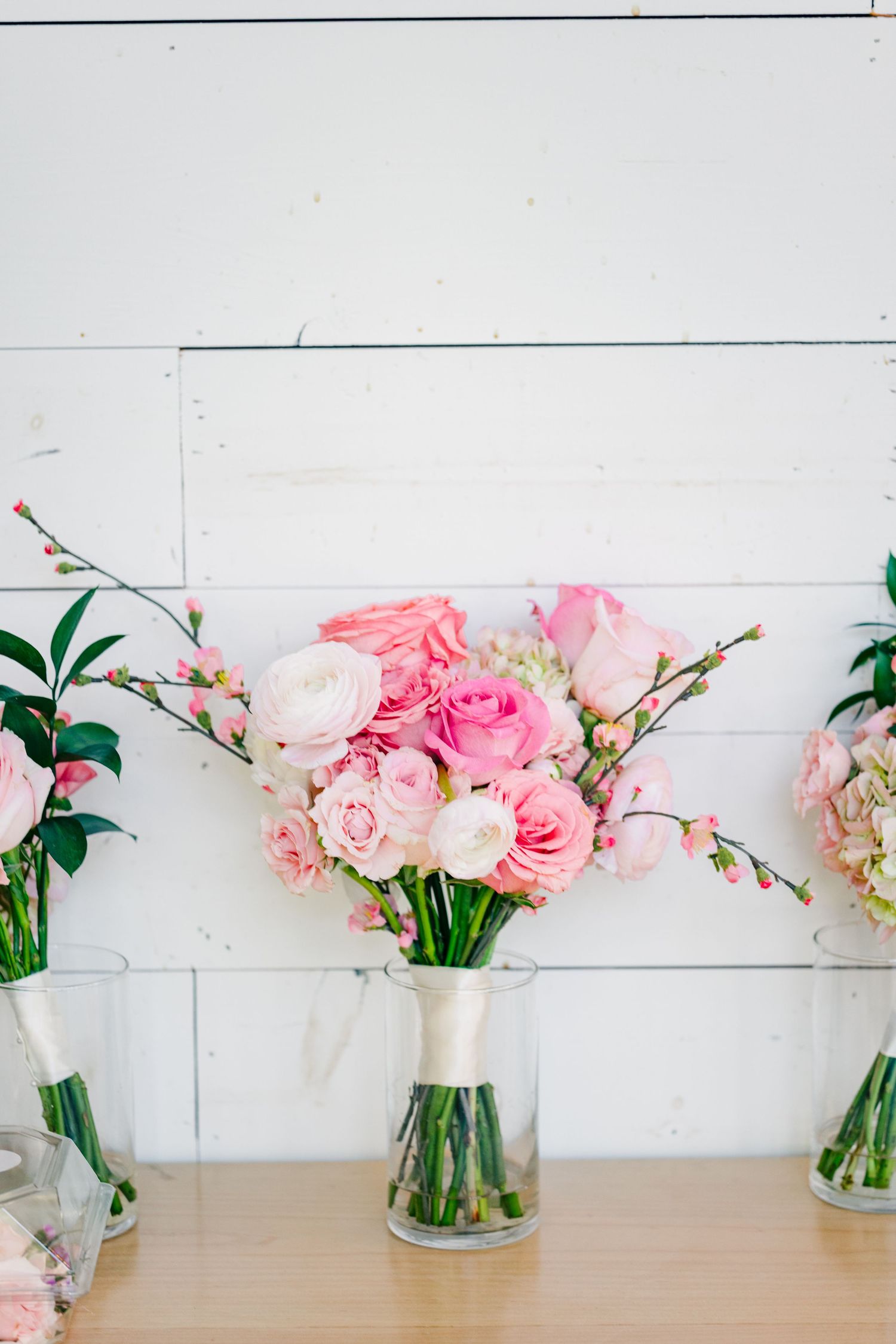 Pink roses and ranunculus bridesmaid bouquets in glass vases displayed against white shiplap wall.