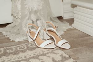 Elegant white bridal sandals with ankle straps sit on a wooden floor beside flowing wedding dress fabric.