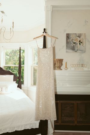 A sparkly white sequin wedding dress hangs on a wooden hanger in a bright bedroom with white decor and floral artwork.