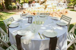 Elegant outdoor wedding reception table set with white linens, place settings and white chairs in a garden setting.