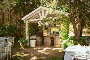An elegant outdoor bar setup with a white pergola, rustic barrels, and scattered dining tables in a garden setting.