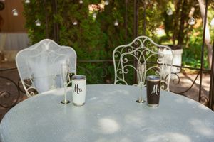 A white metal garden table with ornate chairs and two glasses filled with dark and light beverages in soft sunlight.