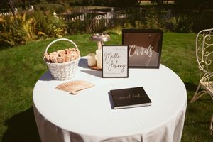 Wedding reception table setup with guest book, cards box, and white wicker basket on round table in garden setting.