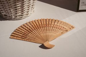 A decorative wooden folding fan with intricate carved patterns lies open on a white surface in natural light.