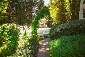 Garden pathway leading through lush greenery to an elegant white wedding gazebo in dappled sunlight.