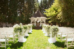 An outdoor wedding ceremony setup with white chairs, floral arrangements, and a gazebo surrounded by trees.