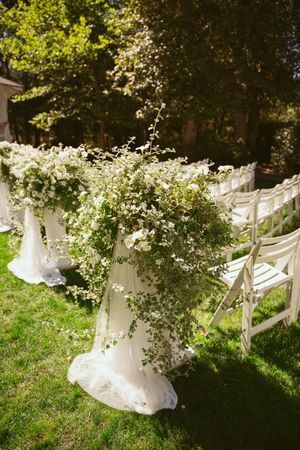 White decorative chairs and floral arrangements line a garden path under lush green trees for an outdoor wedding ceremony.