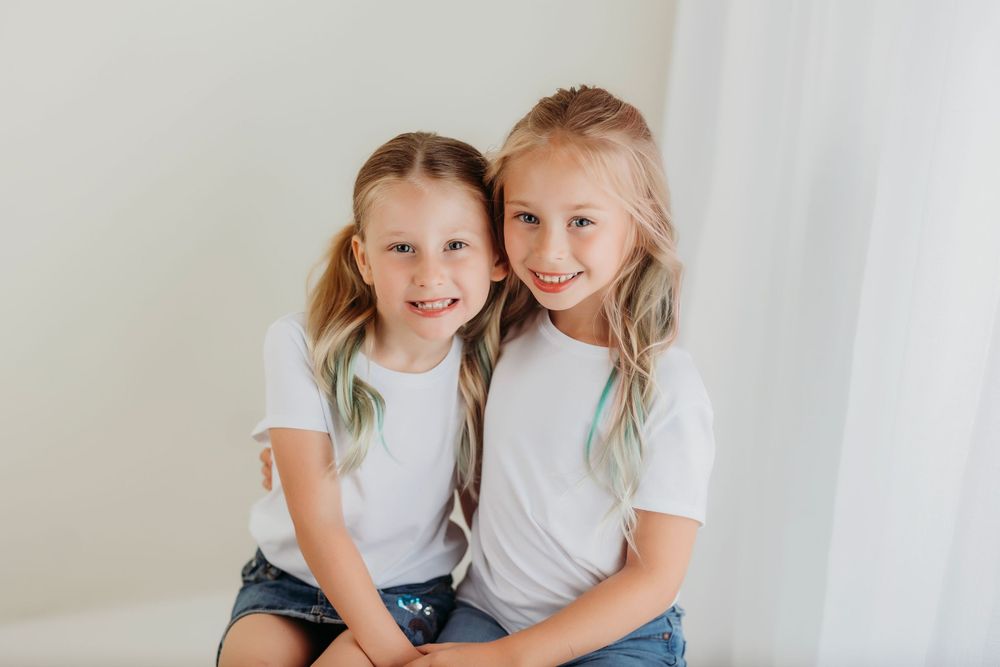 Two young children wearing white shirts share laughter while sitting on a white platform in a minimalist studio setting.