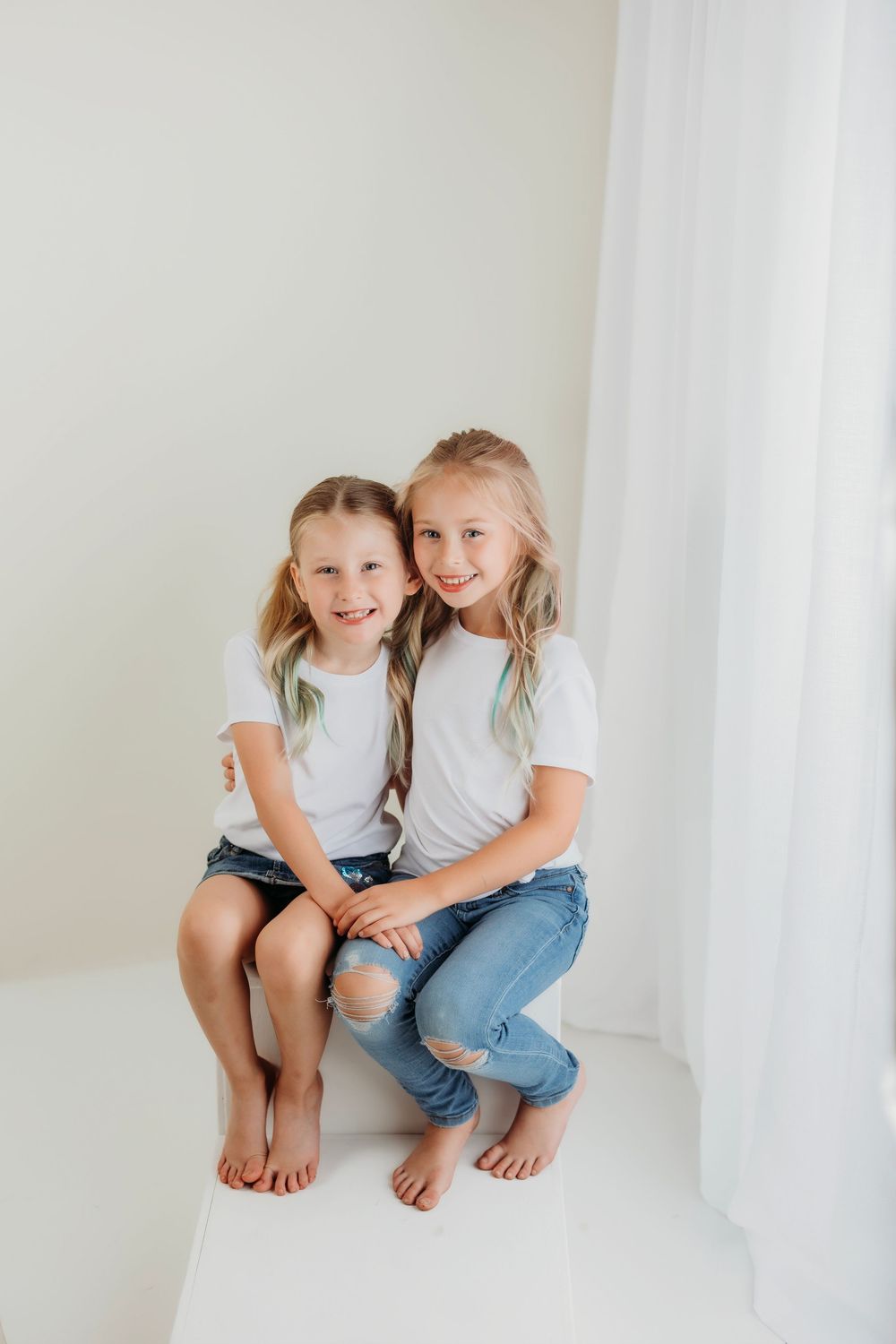 Two kids in casual outfits sit together on a white platform, sharing a playful moment.