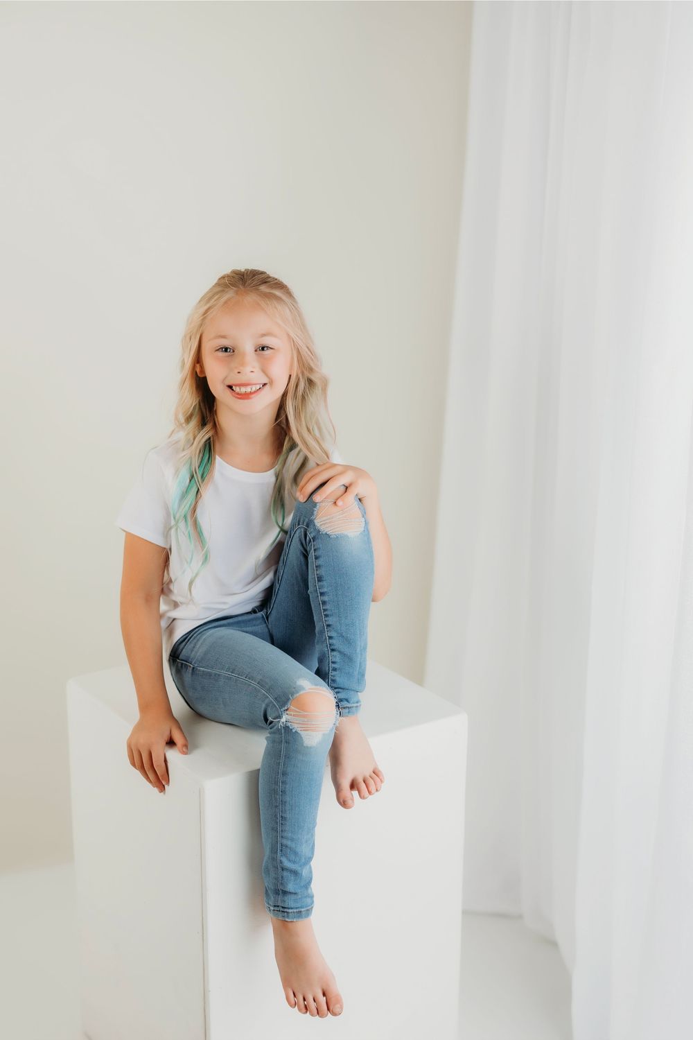 Child in denim and white t-shirt sits casually on white platform in bright studio setting.