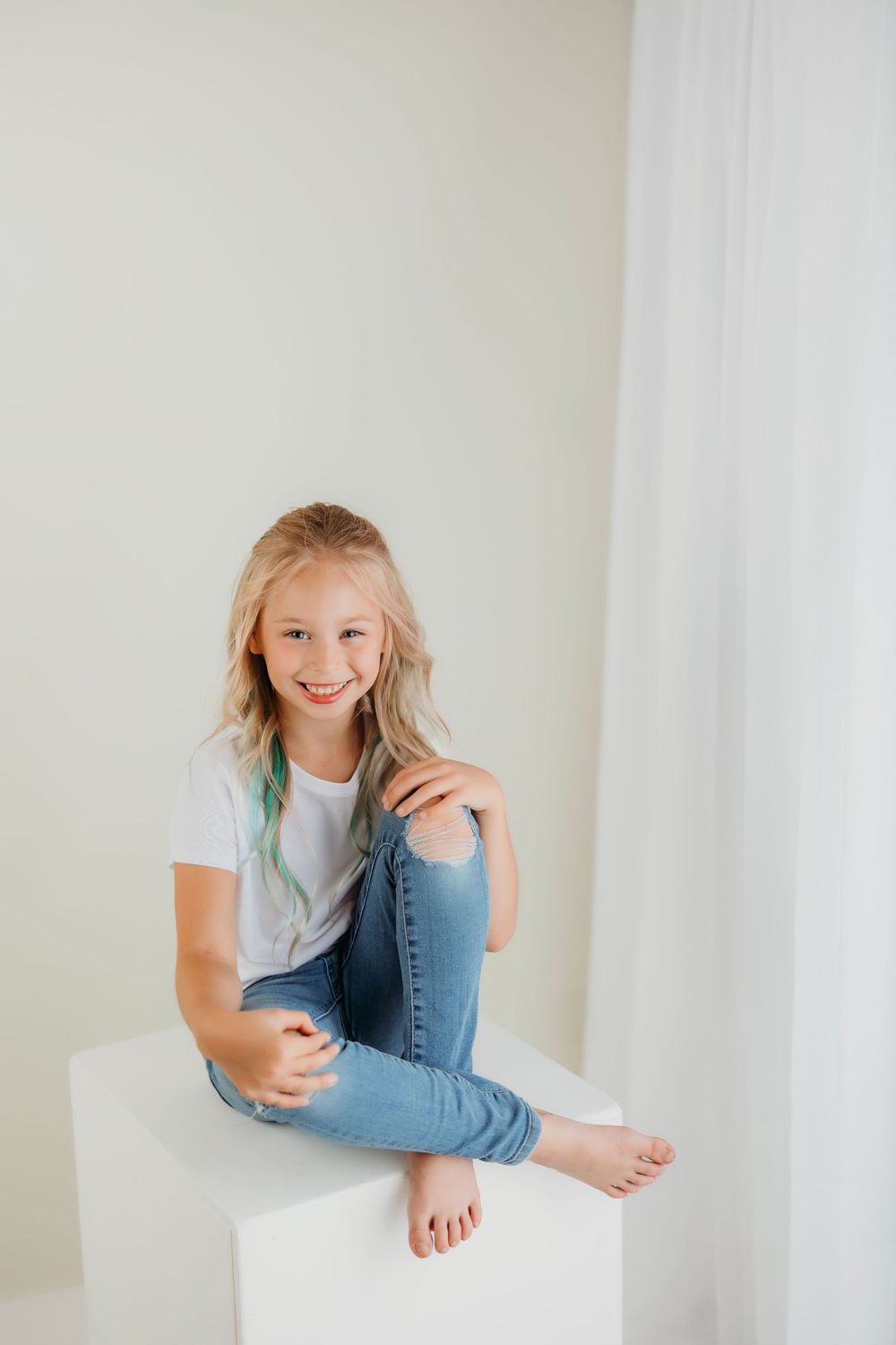 Young person in blue jeans and white tee poses casually on white platform with bright smile.
