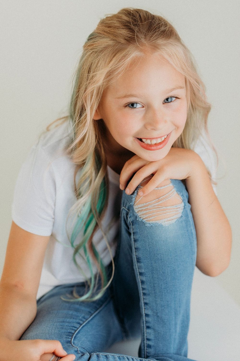 Child with blonde hair and turquoise streak poses playfully in studio setting.