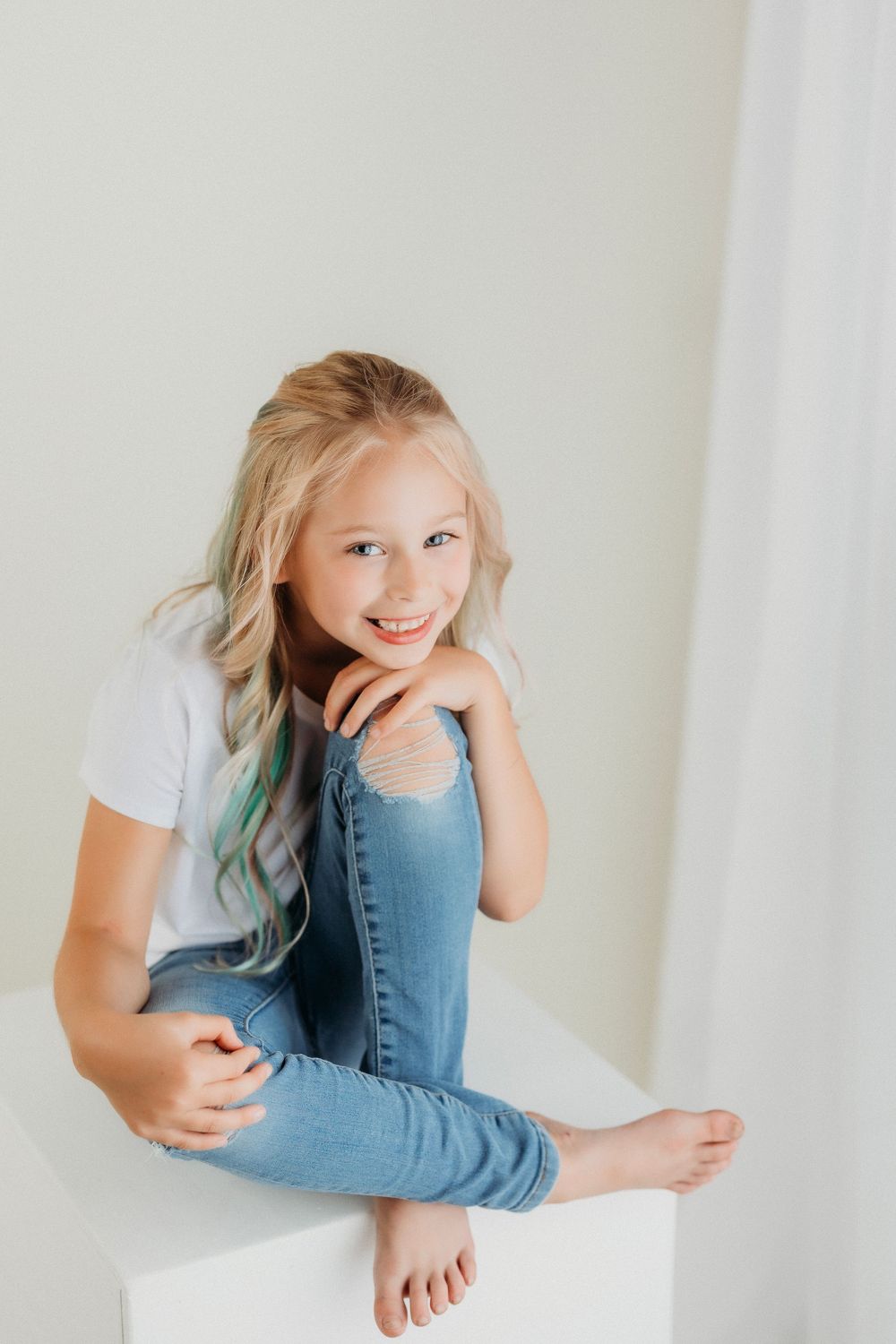 Child in casual attire strikes playful poses while sitting on white platform in studio.