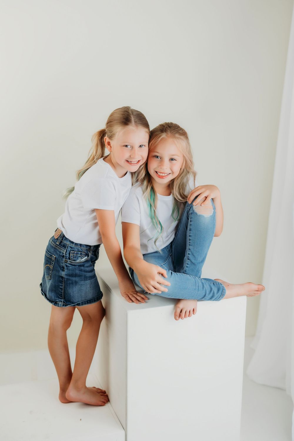 Two people in casual denim and white tops pose playfully against a white wall and cube.