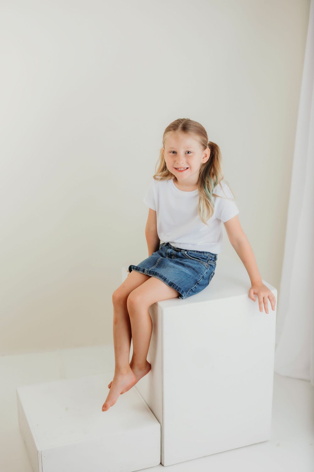 A young person in denim shorts and white t-shirt sits casually on a white cube.