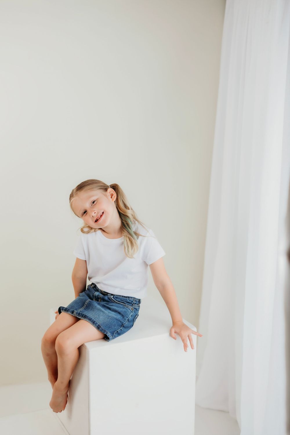 A young person in denim shorts and white shirt poses expressively on a white cube.