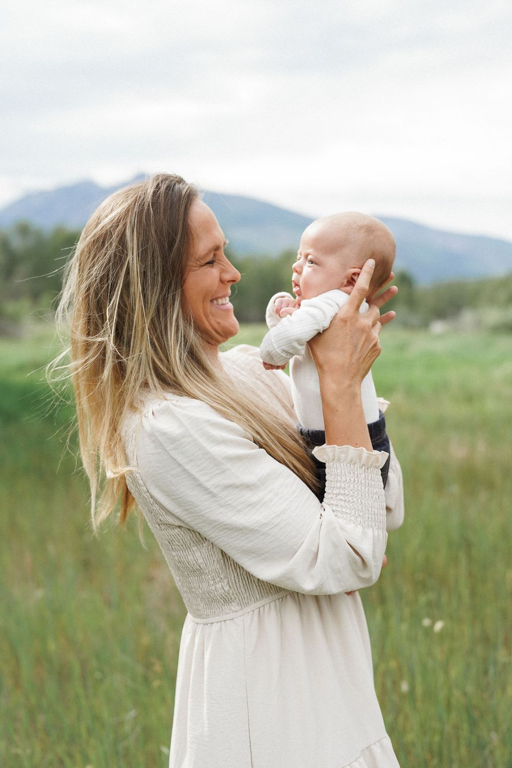 A woman in a white dress holds up a smiling baby against a scenic mountain backdrop in a grassy meadow.