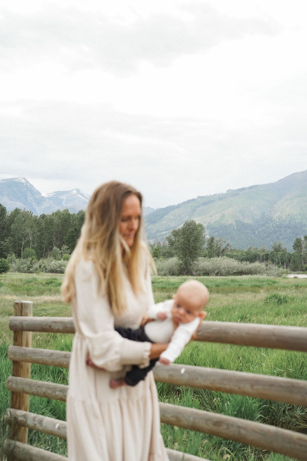 A figure in a white dress poses thoughtfully against a rustic wooden fence with scenic mountain views in the background.