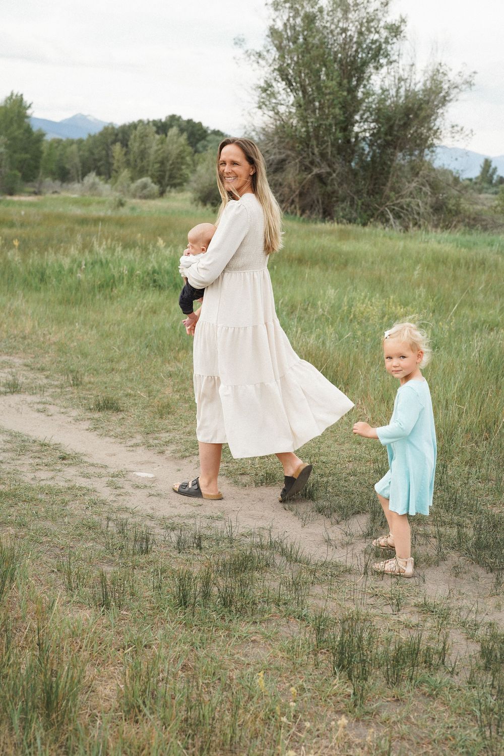 Mother and child walking together on a dirt path through meadow grass wearing white and blue summer outfits.