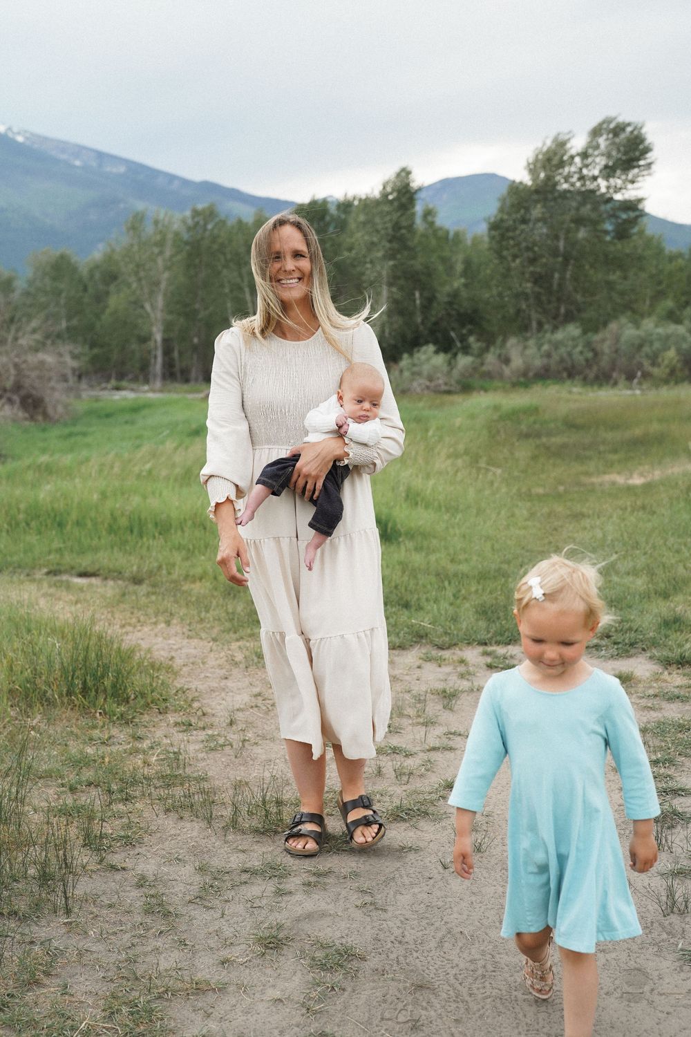 A family walks along a dirt path with mountains and trees in the background on a serene summer day.