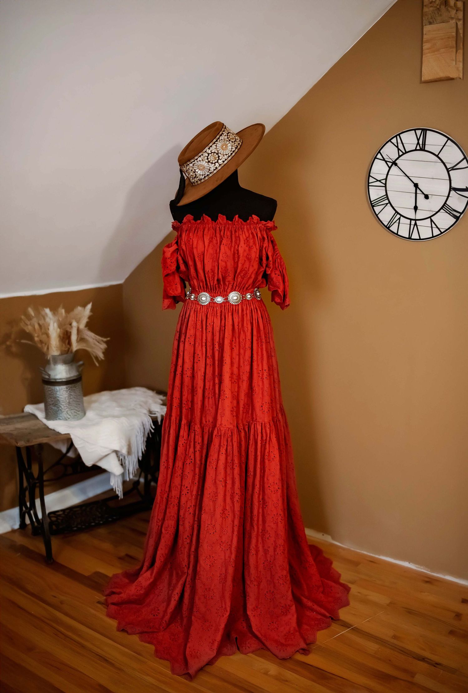 A stunning red off-shoulder maxi dress with lace details and brown hat displayed on mannequin against beige wall with clock.