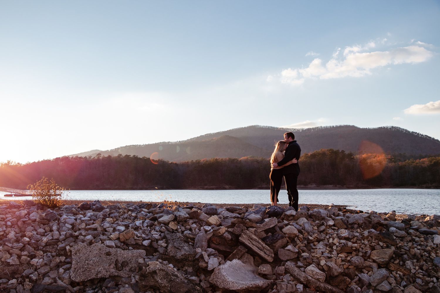 Couple embraces by a lake at sunset, standing on rocky ground with soft light and mountains behind them.
