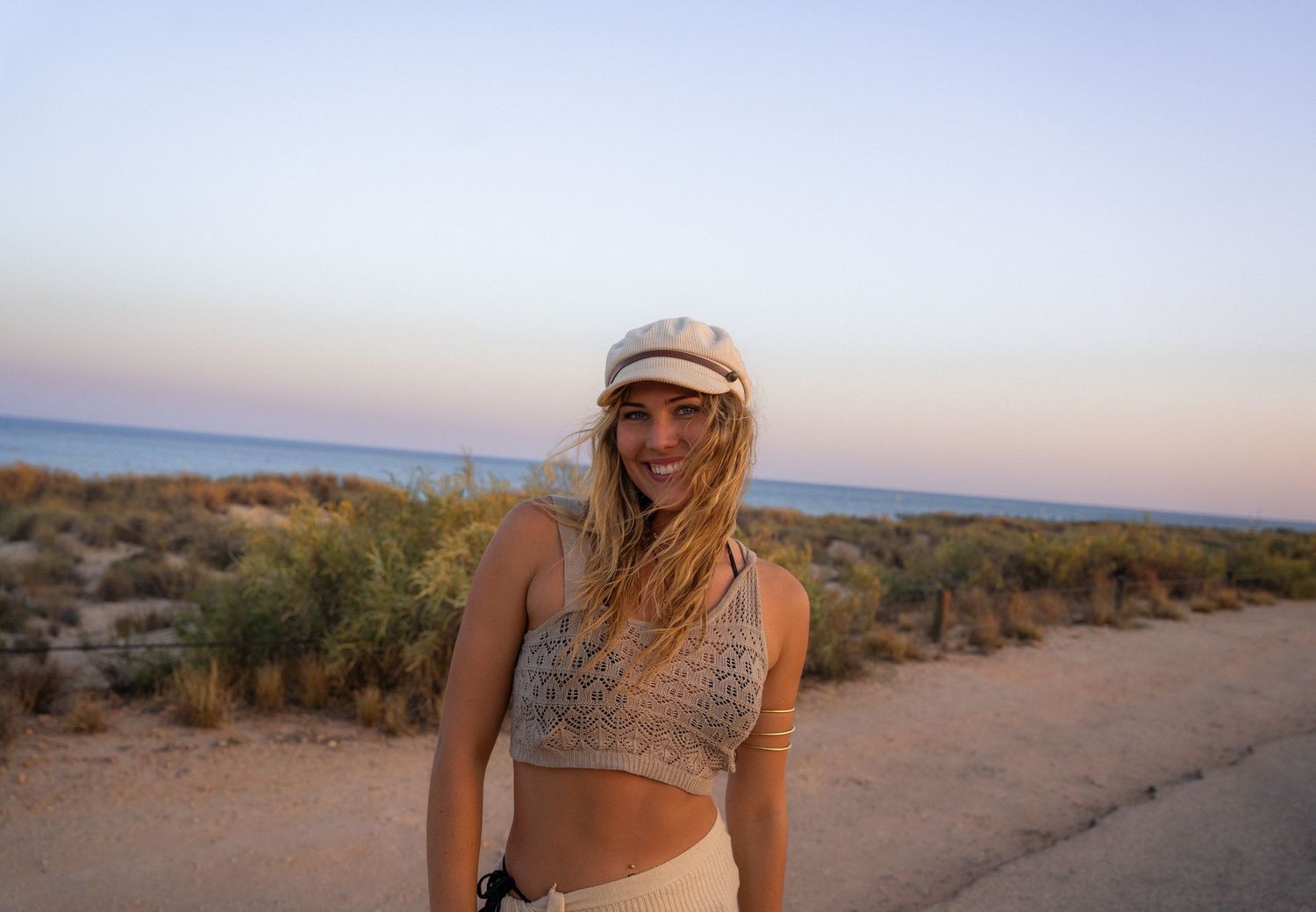 Woman in crop top and cap poses against desert landscape at sunset.