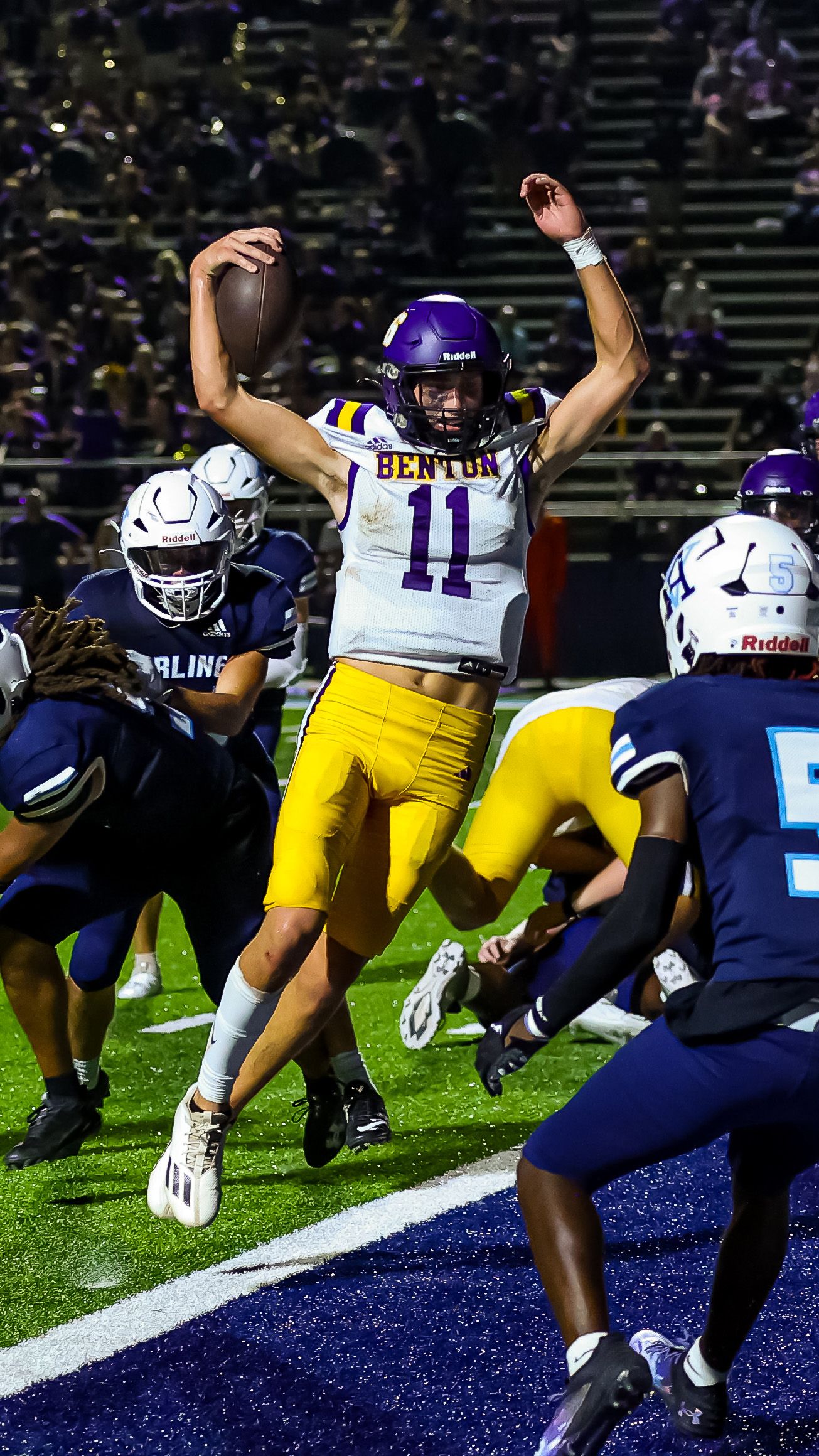 Football player in purple and yellow Benton uniform leaps in the air during game action against opposing team.
