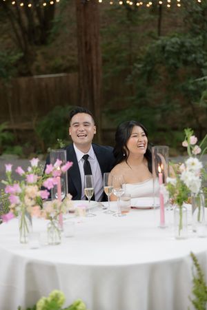 A couple enjoys their outdoor wedding reception at a table with pink floral centerpieces and string lights overhead.