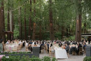 An outdoor wedding reception in a wooded forest setting with string lights and guests seated at tables among tall trees.